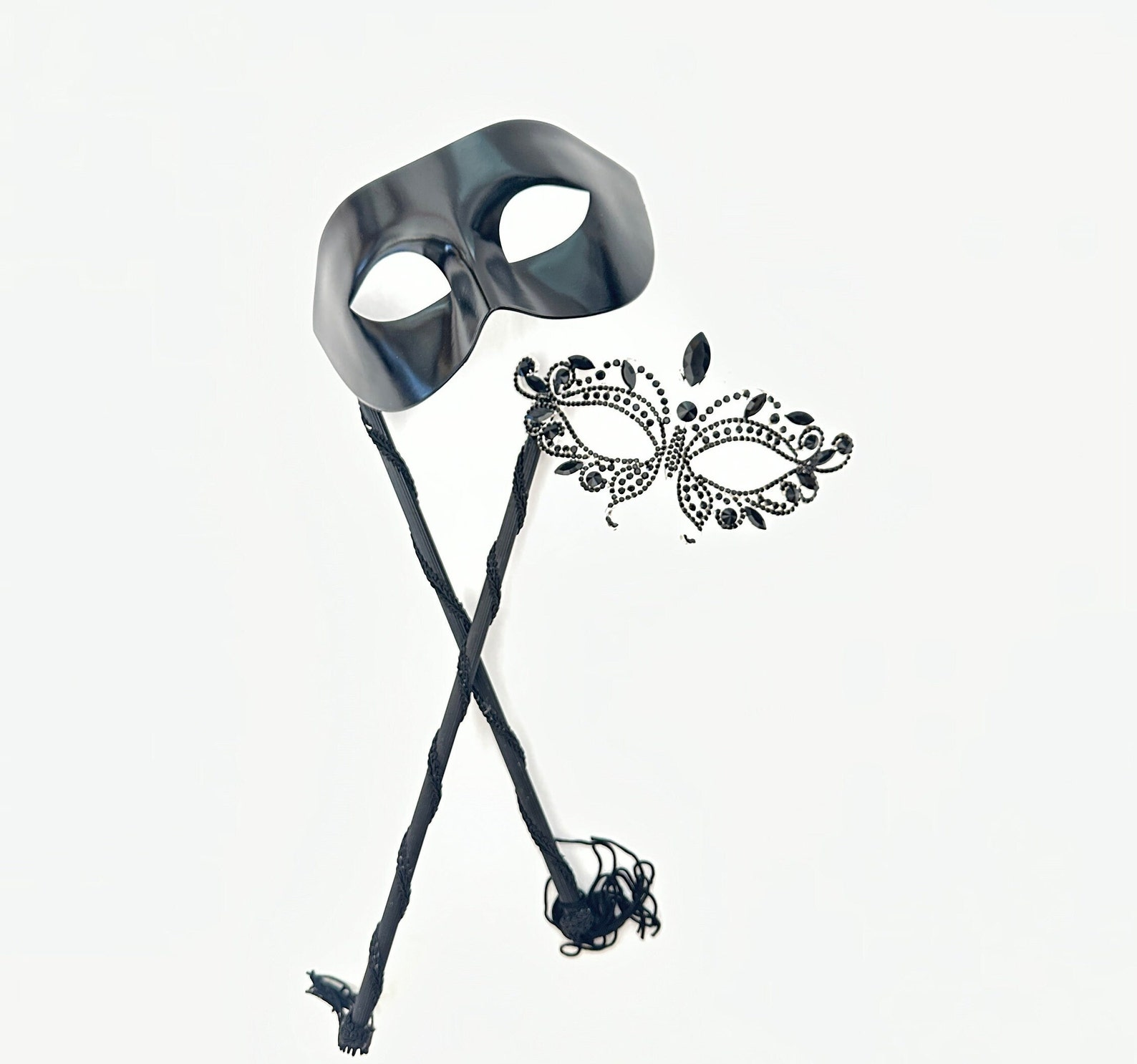 Black phantom-style men’s masquerade mask on stick with matching women’s rhinestone filigree mask, designed for elegant couple’s wear.