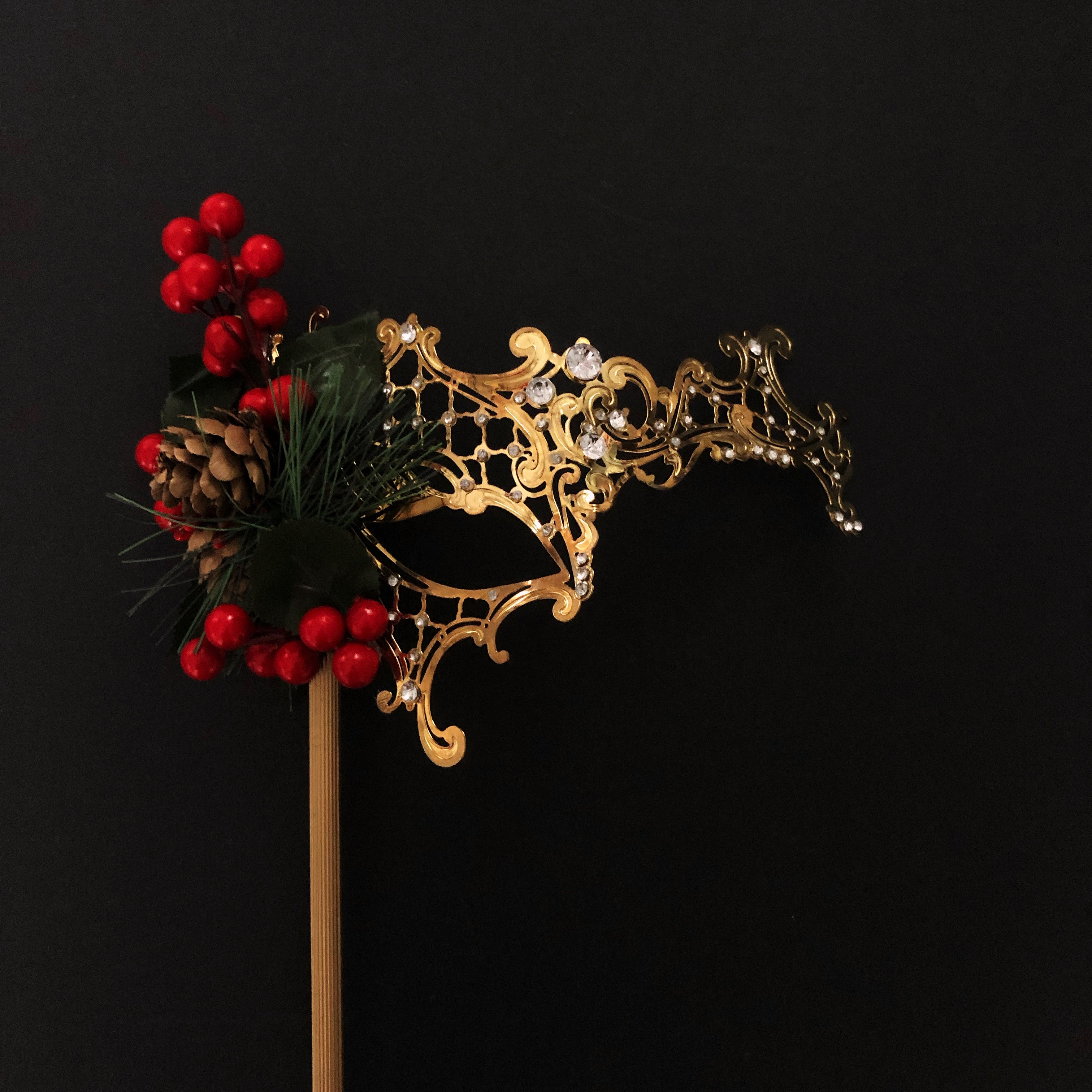 Gold filigree Christmas masquerade mask with holly berries, pinecone, and evergreen accents on a stick.