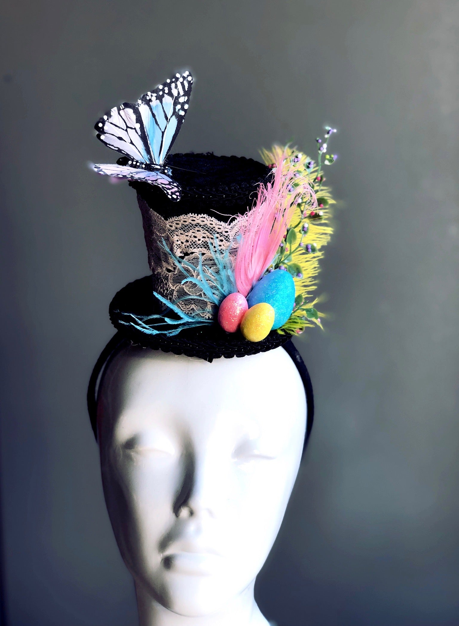Easter themed fascinator with a miniature top hat decorated with Easter eggs, pastel feathers, a butterfly in flight and floral accents attached on a headband