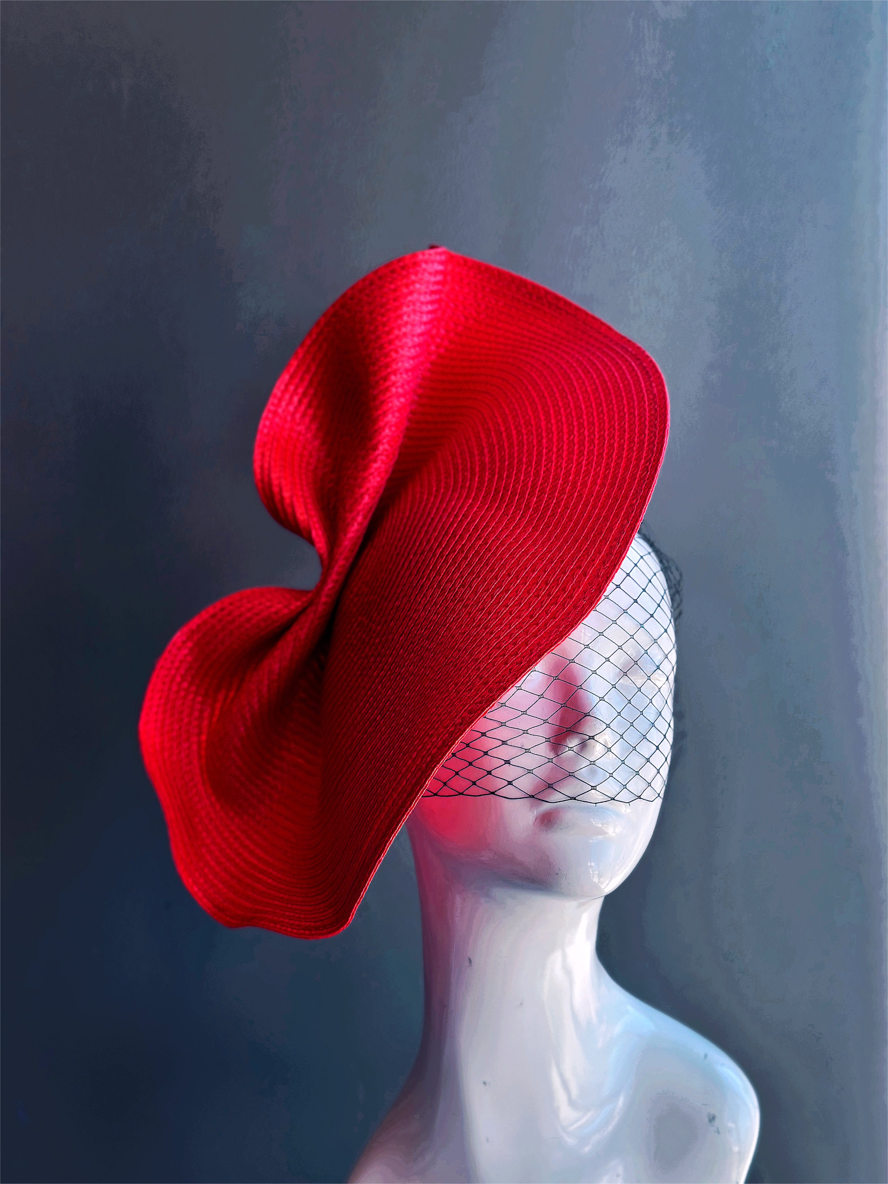 Crimson red sculptural fascinator hat with modern twist and black mesh face veil for derby or formal event wear