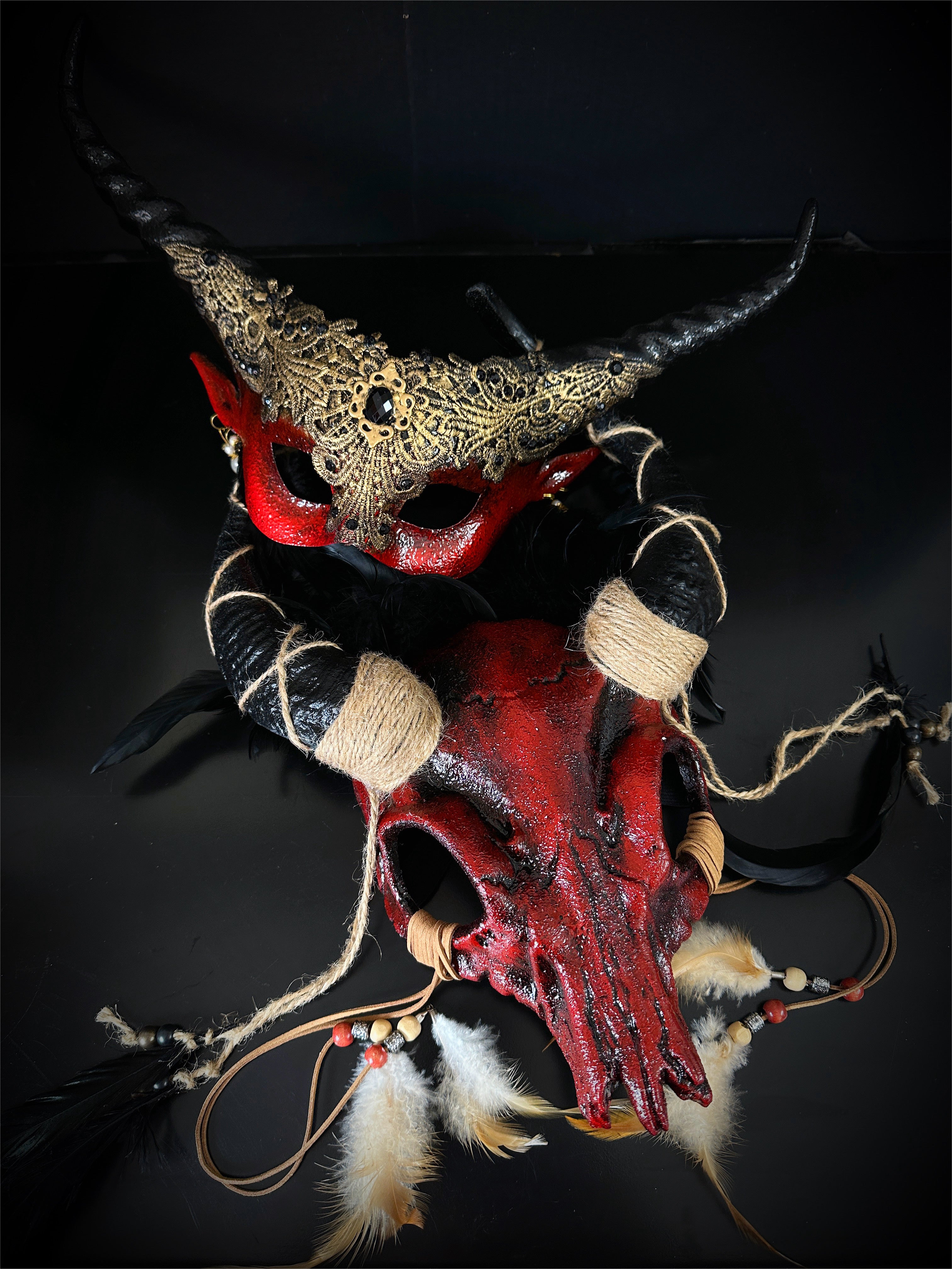 Red skull headdress with feathers and rope details paired with black and gold filigree horn masquerade mask