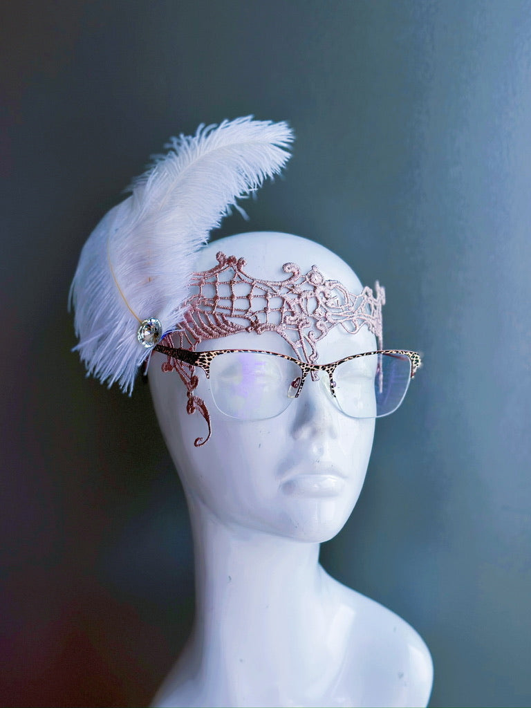 Elegant lace masquerade mask in rose gold worn with glasses, featuring a large white feather accent