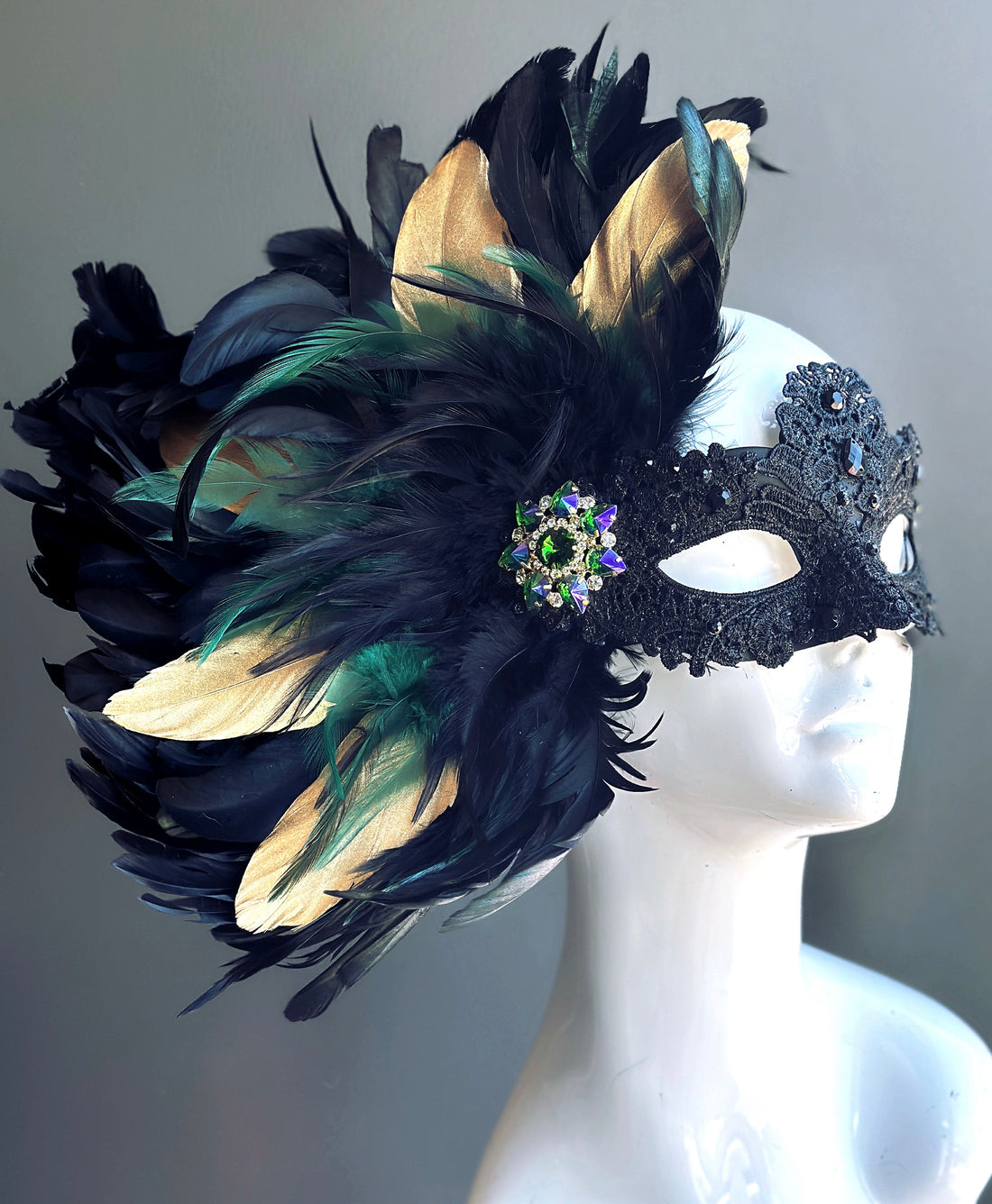 Black lace masquerade mask for women with gold and green feathers and emerald brooch.