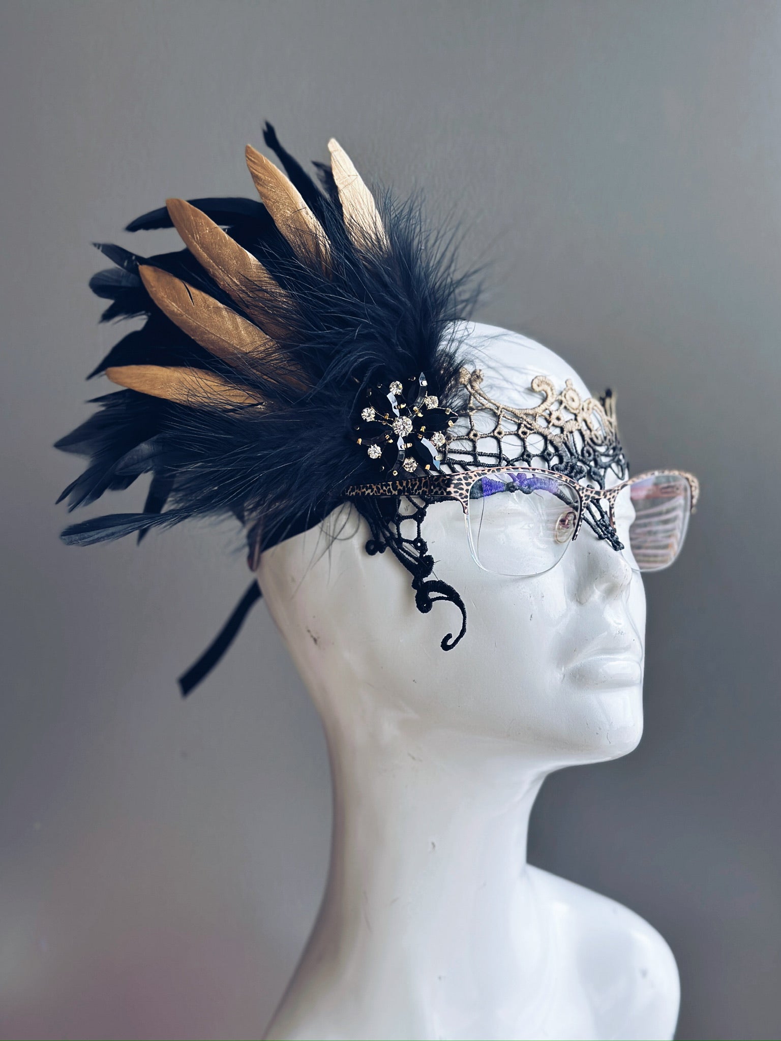 Gold lace eyeglass mask with black and gold feathers, rhinestone embellishments, and an elegant filigree design.