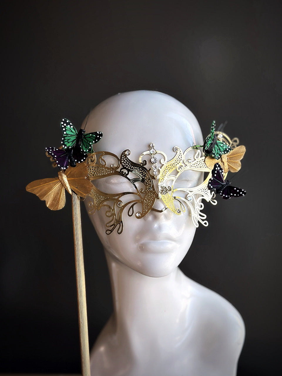 Gold handheld masquerade mask with intricate filigree and butterfly accents displayed on a mannequin head.