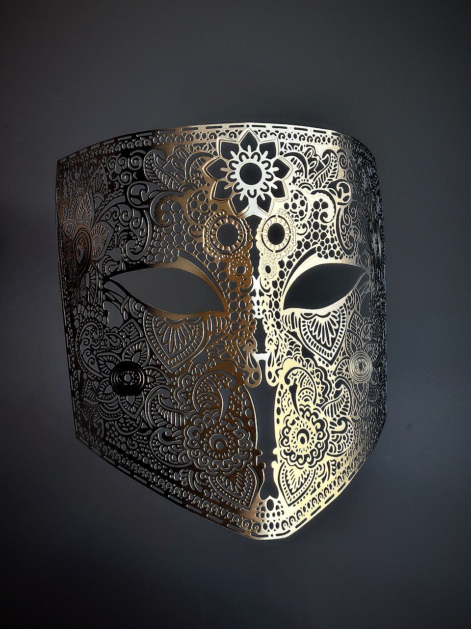 Ombre full-face mask with intricate laser-cut filigree, fading from gold in the center to black on the sides, set against a dark background.