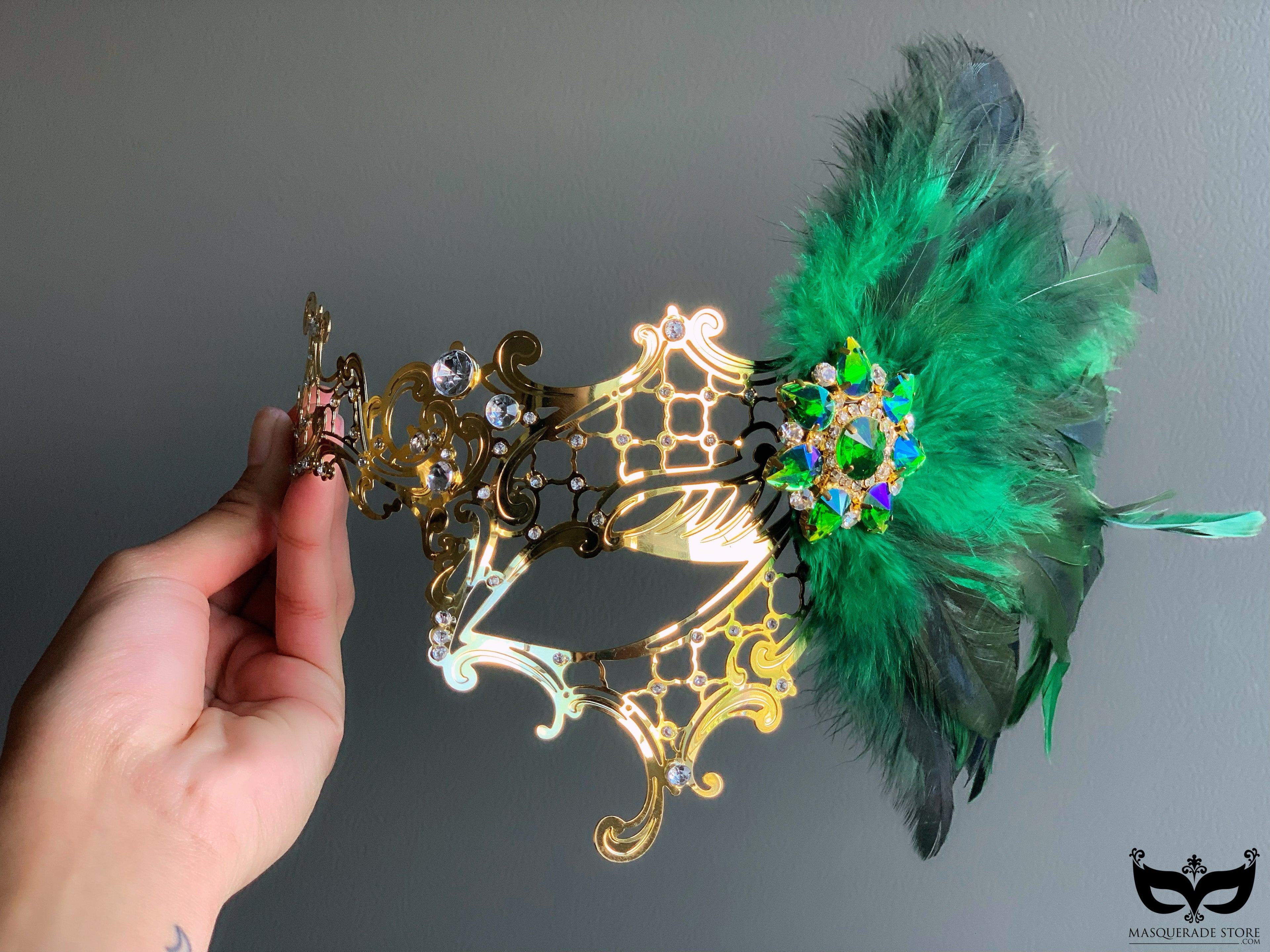 Gold metal filigree mask adorned with green feathers and a multicolored brooch, ideal for luxury events and masquerade balls.