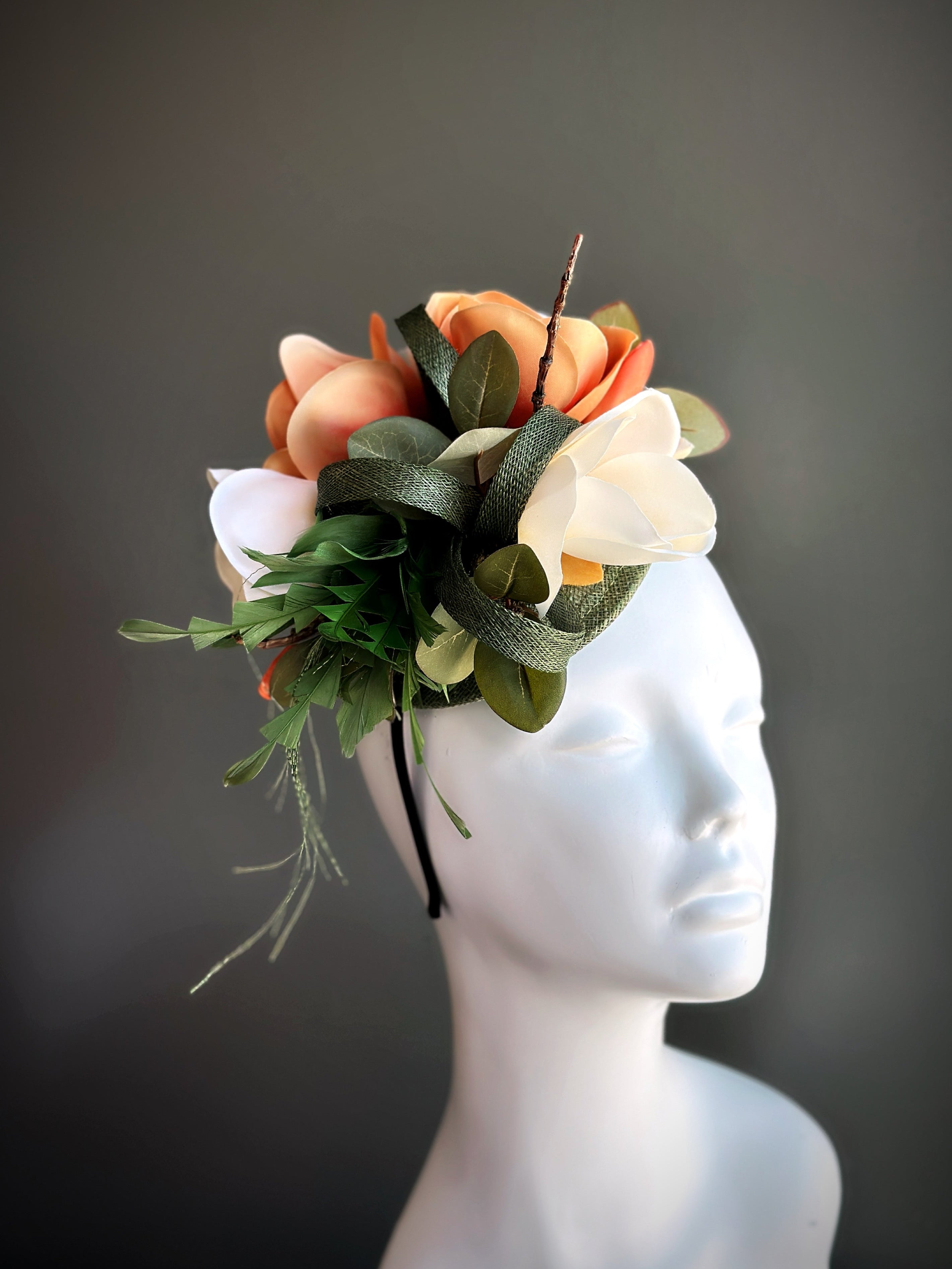 Fascinator hat with ivory and peach magnolia flowers, accented by greenery and elegant ribbon for spring or summer occasions.