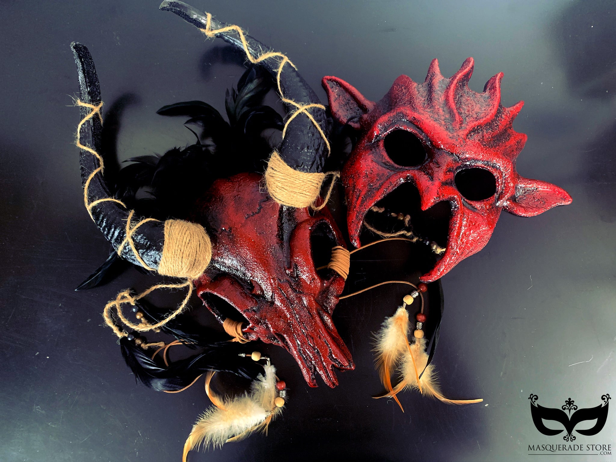 A set of two masks: a devilish red demon mask with horns and a rustic ram mask decorated with feathers and twine, ideal for Halloween or cosplay.