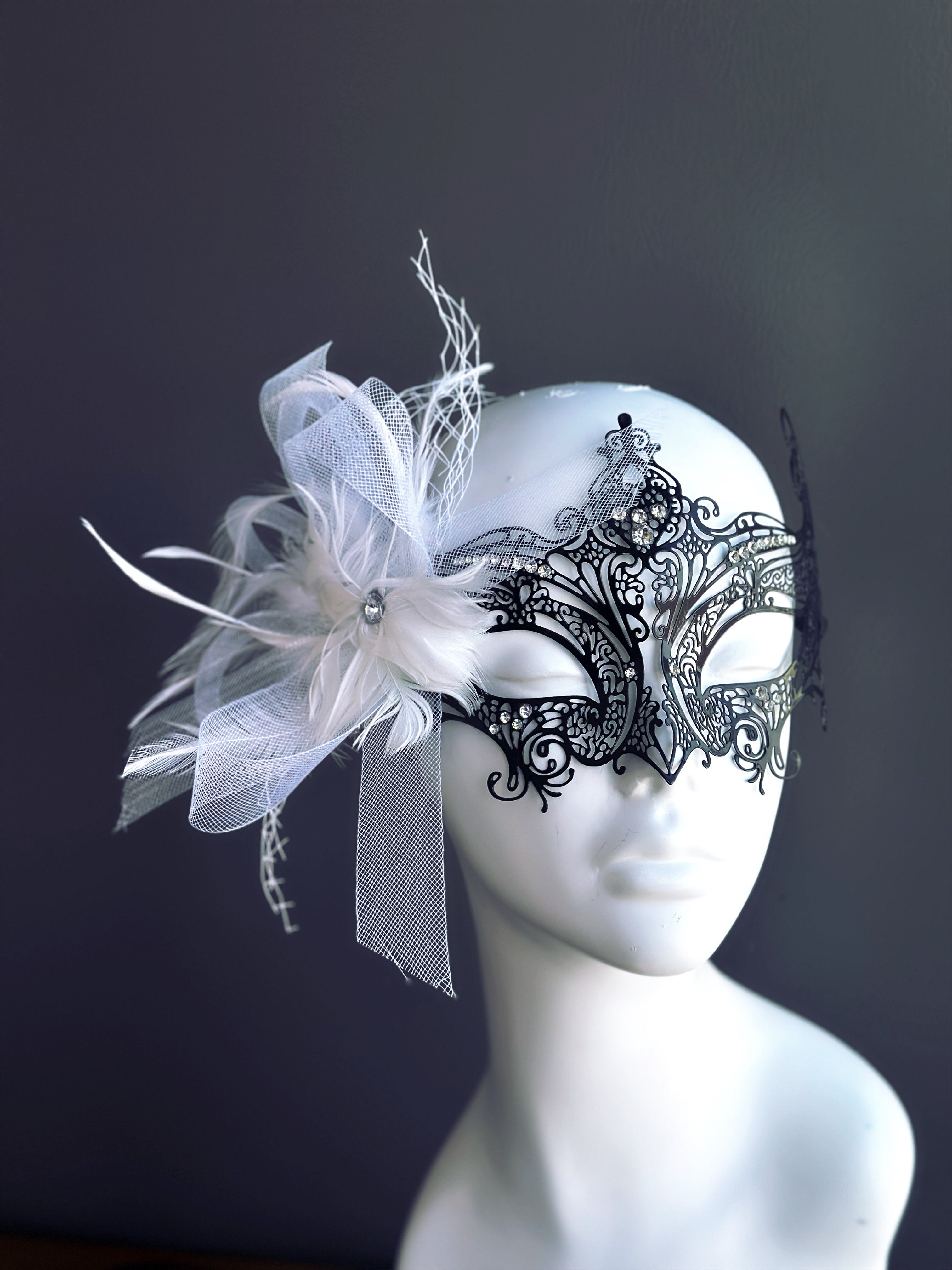 Black metal masquerade mask with rhinestones and white feathers on mannequin