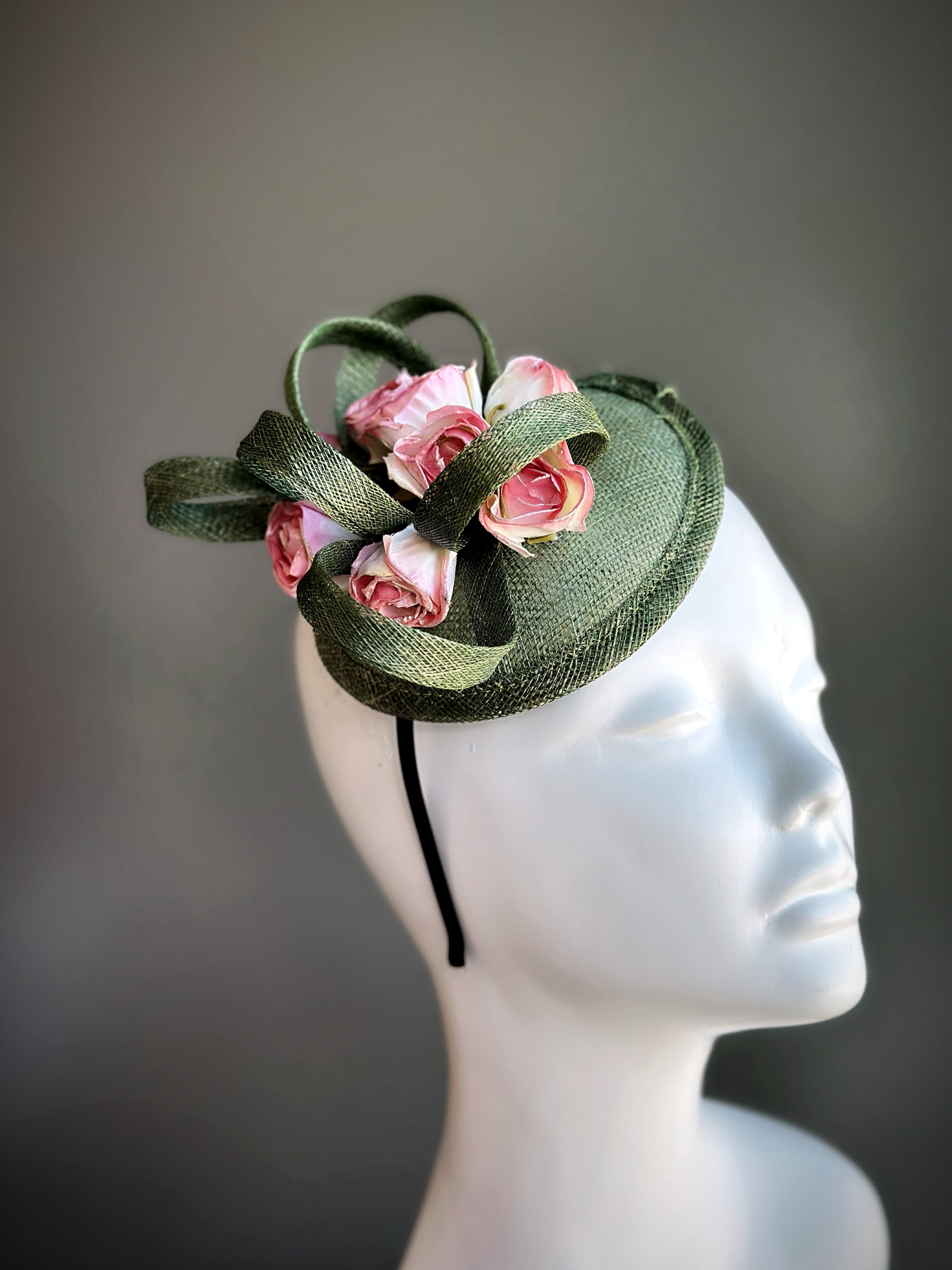 Green fascinator hat with sculpted ribbon design and soft pink roses, ideal for spring garden parties or weddings.