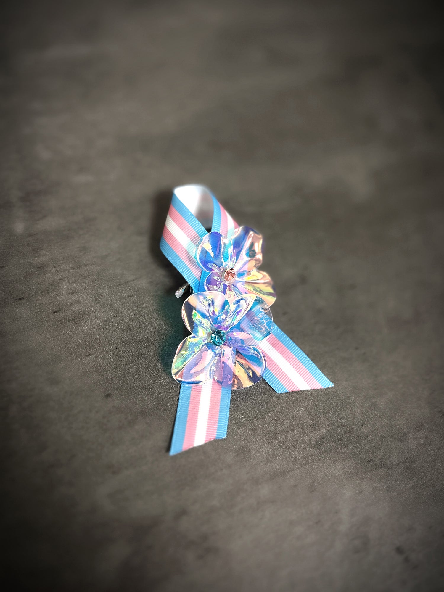 Transgender pride ribbon pin with iridescent flower embellishments on pink, white, and blue stripes