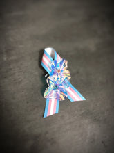 Transgender pride ribbon pin with iridescent flower embellishments on pink, white, and blue stripes