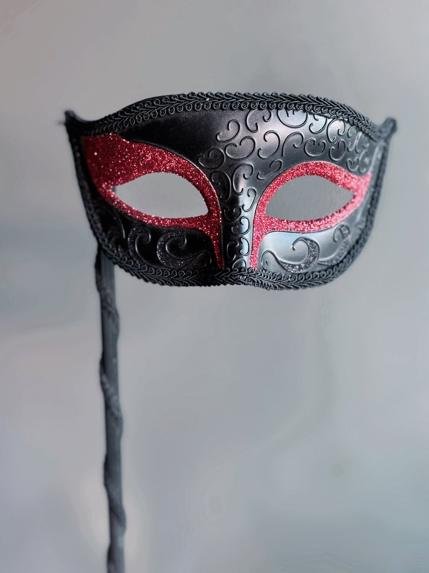 Black handheld masquerade mask with red glitter accents and ornate swirl details