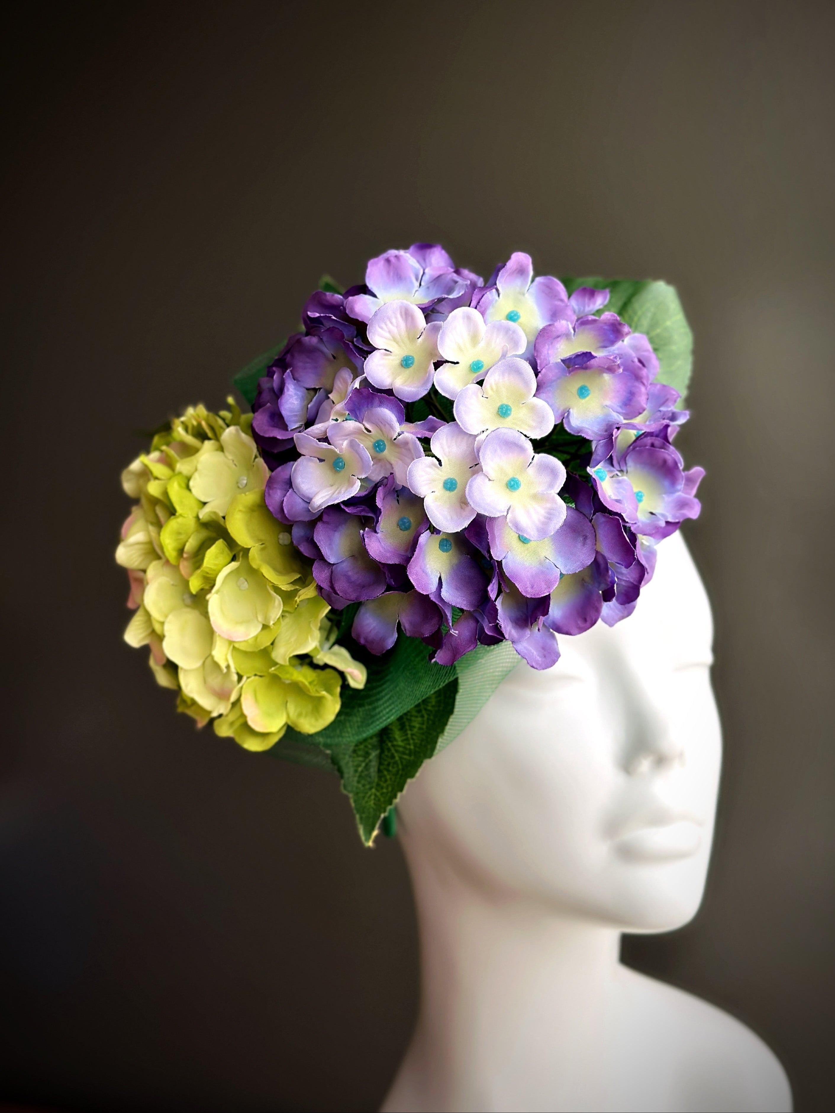 Fascinator hat with purple and green hydrangea flowers and green headband on mannequin.