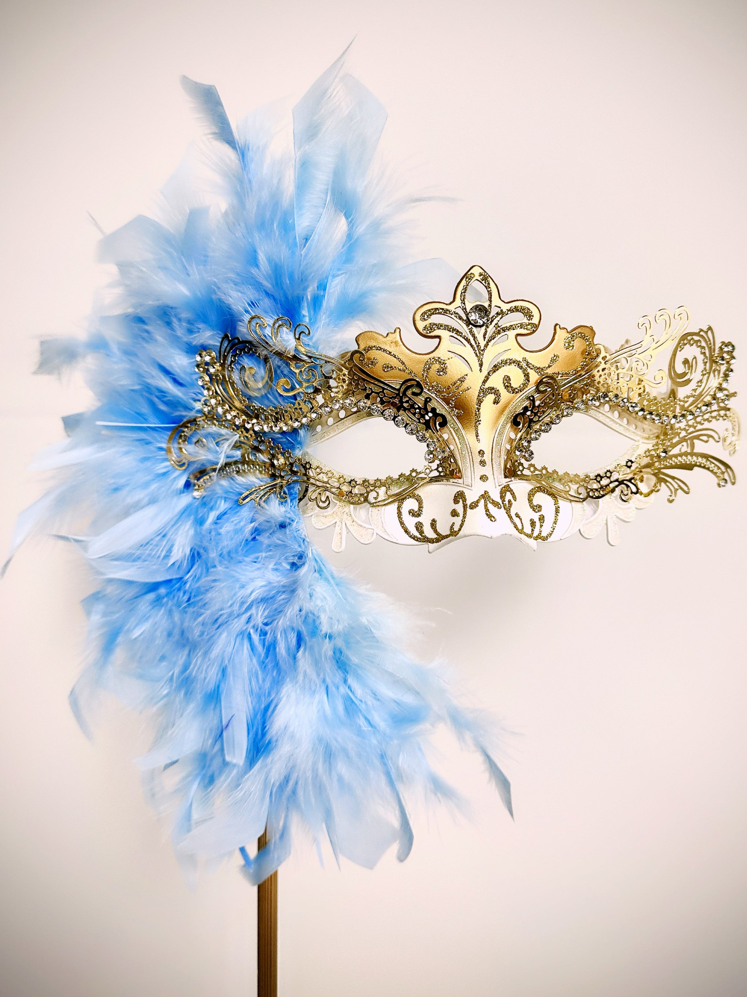 Gold white masquerade mask on a gold stick with light blue feathers.