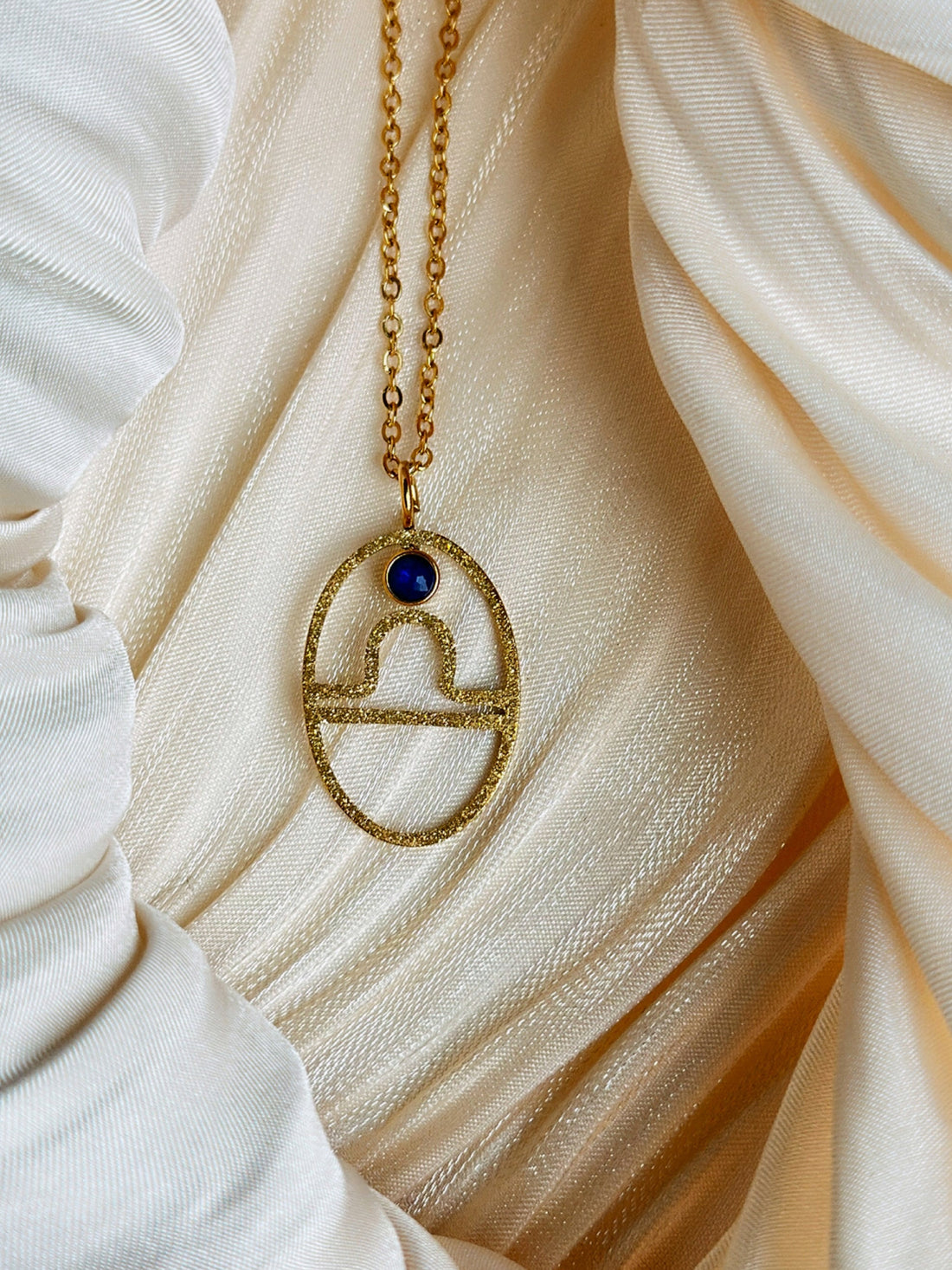 Gold Libra Zodiac necklace with a blue gemstone pendant on a white fabric background