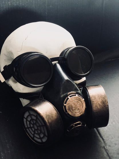 Black and antique bronze cosplay gas mask with dark round goggles, dual filter canisters, adjustable straps, and industrial dystopian styling.