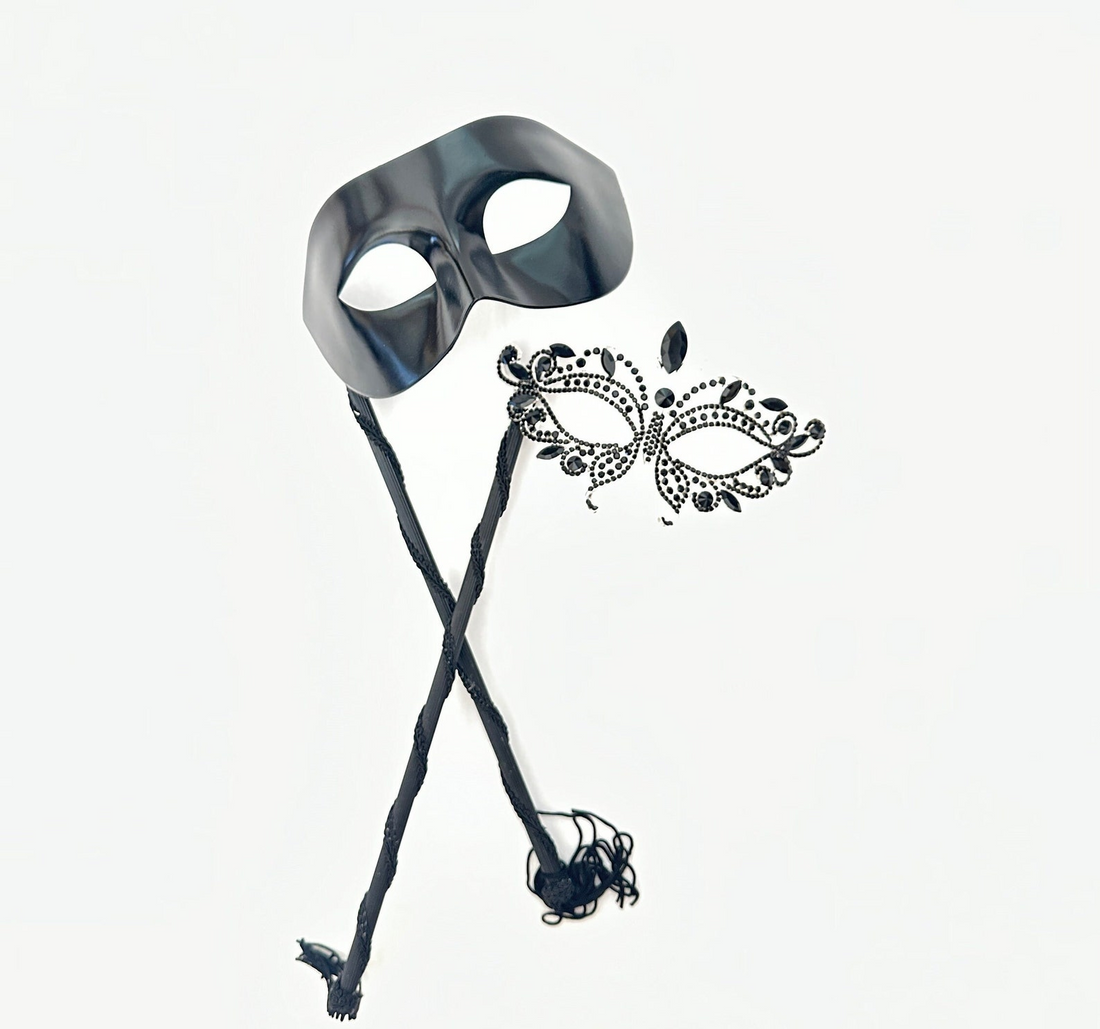Black phantom-style men’s masquerade mask on stick with matching women’s rhinestone filigree mask, designed for elegant couple’s wear.