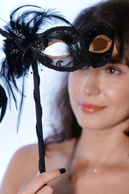 Woman holding a handheld black masquerade mask with feathers and lace trim.