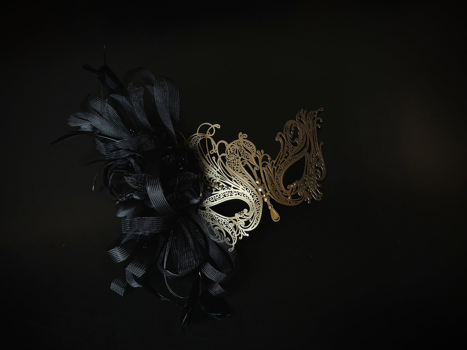 Black and gold masquerade mask with filigree metal, tulle ribbon, and feathers, designed for proms and costume balls