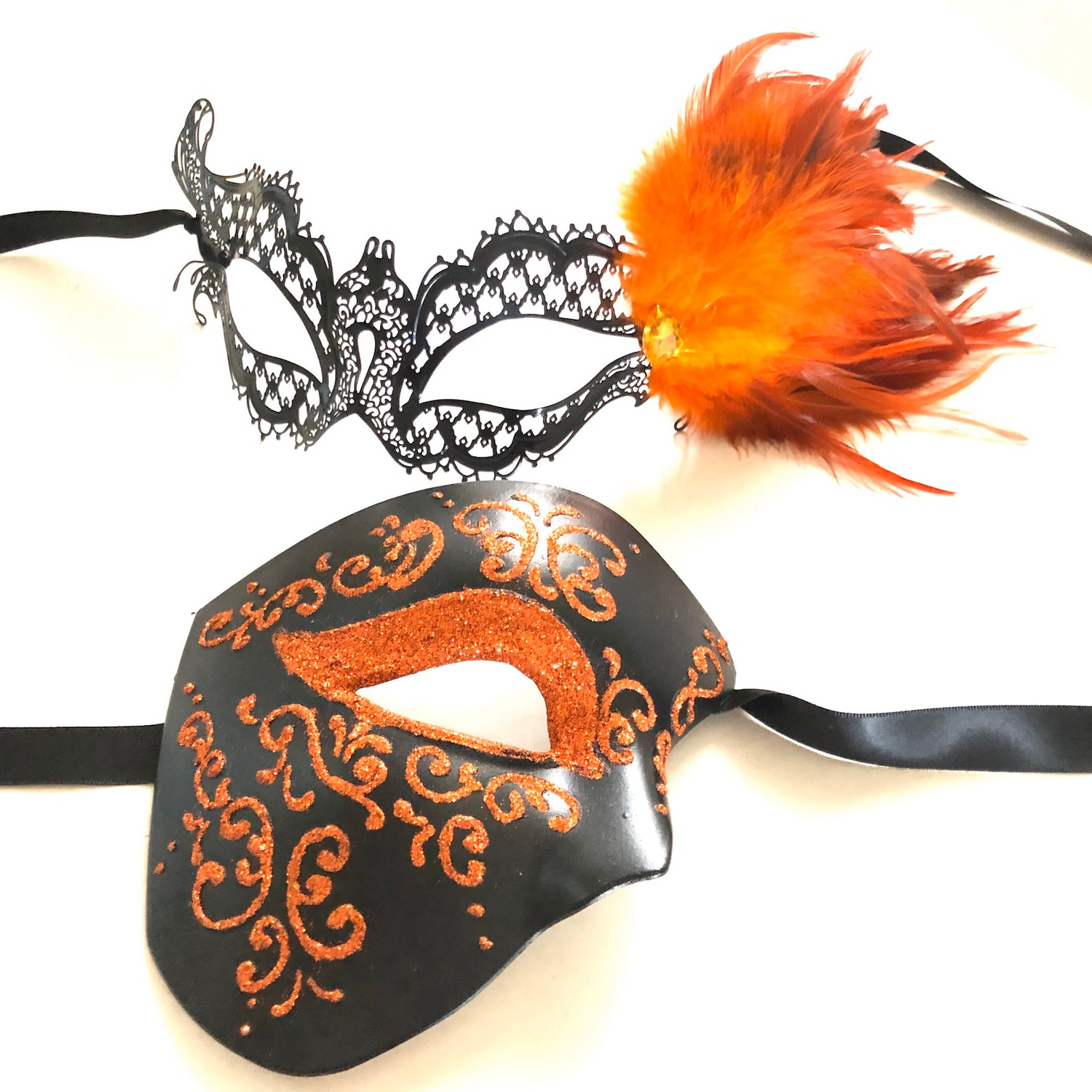 Black and orange couples masquerade mask set featuring men’s black mask with orange glitter scrolls and women’s filigree mask with orange feathers.