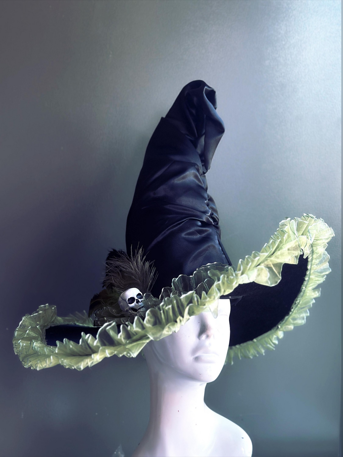 Black witch hat with skull accent, green ruffle brim, and feather detail.