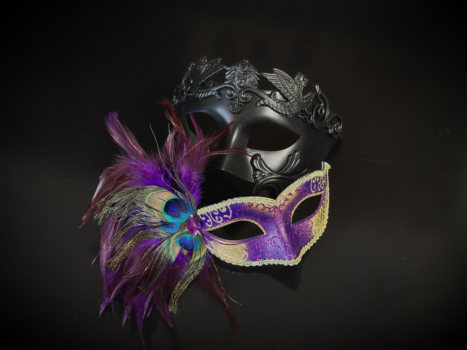 Two masks, one with feathers and the other black with intricate design, on a dark background.