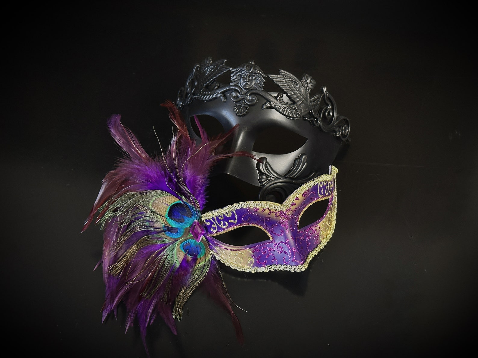 Two masks, one with feathers and the other black with intricate design, on a dark background.