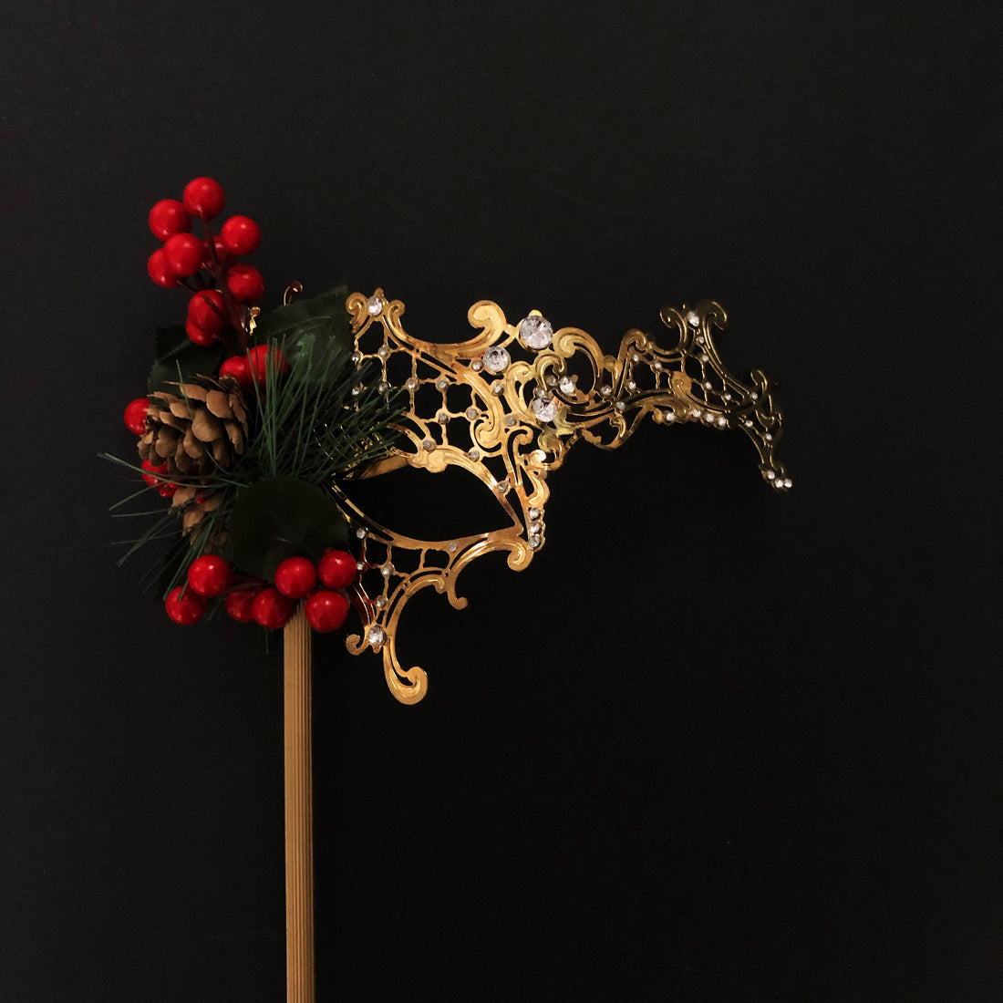 Gold filigree Christmas masquerade mask with holly berries, pinecone, and evergreen accents on a stick.