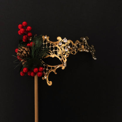 Gold filigree Christmas masquerade mask with holly berries, pinecone, and evergreen accents on a stick.