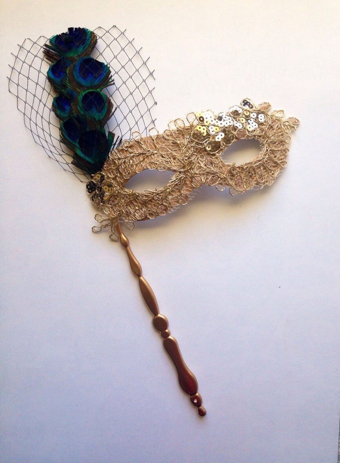 Gold lace masquerade mask on stick with sequins, peacock feathers, and net accent.