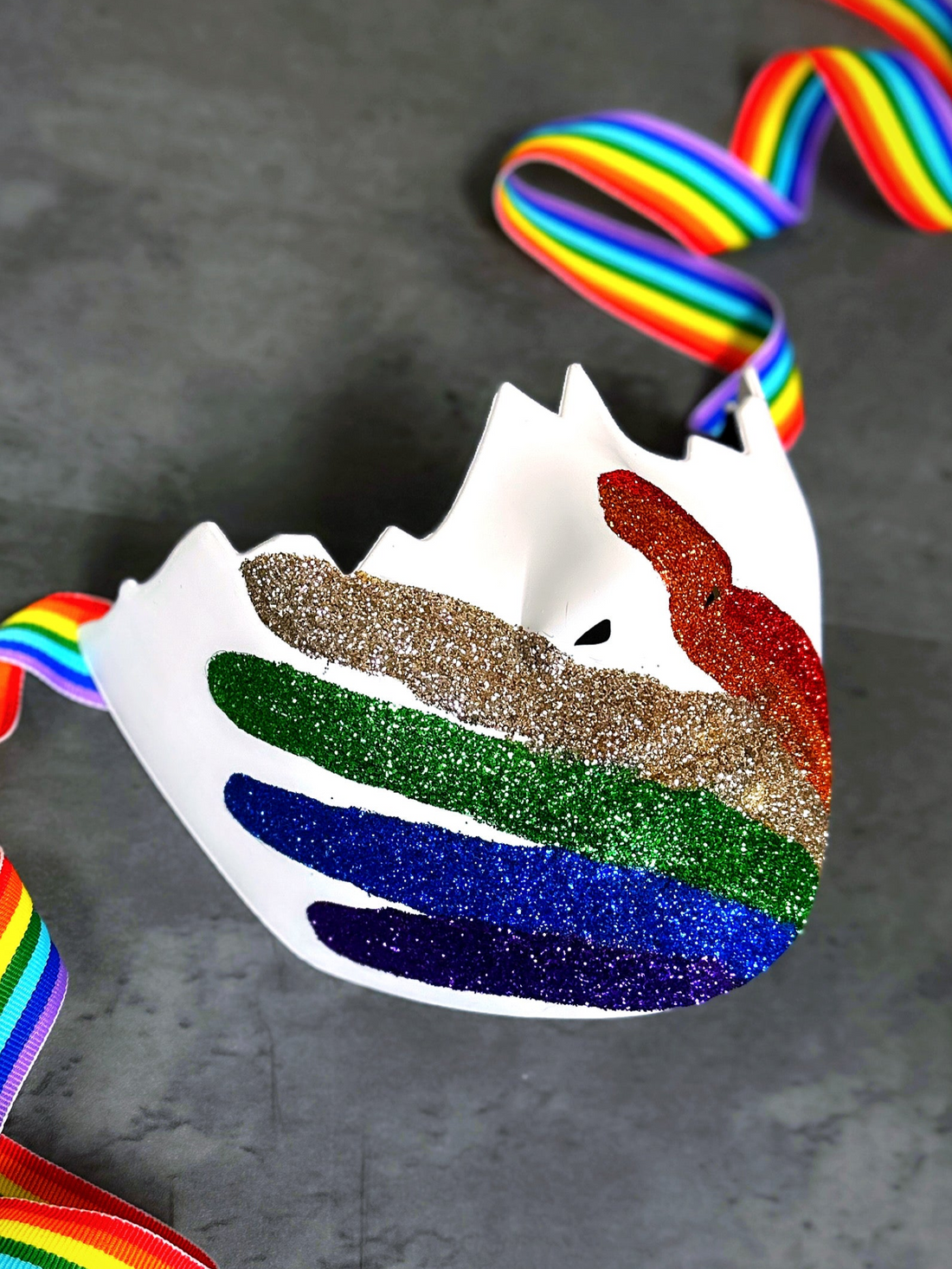Pride glitter mask featuring rainbow handprint design and satin rainbow straps on gray background

