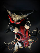 Red skull headdress with feathers and rope details paired with black and gold filigree horn masquerade mask