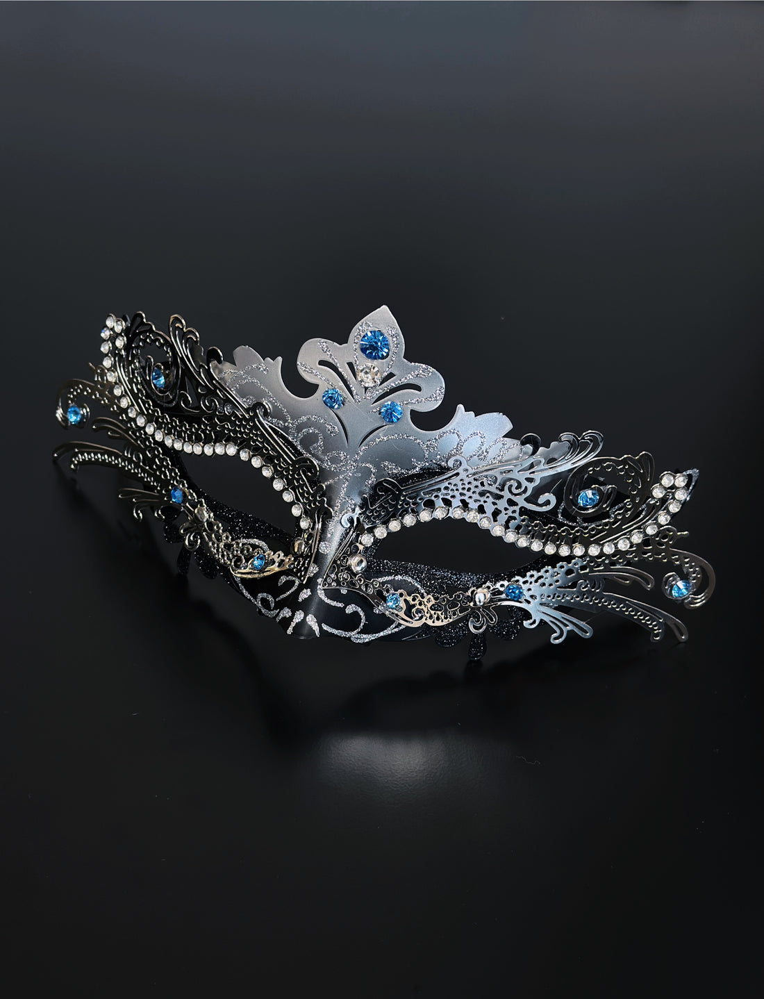Silver and black women’s masquerade mask with filigree design, glitter accents, and blue rhinestones, ideal for Venetian masquerade balls and parties.