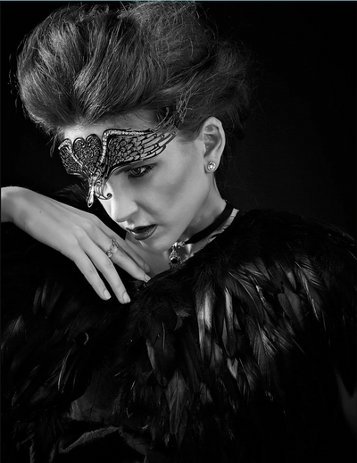 Metal masquerade mask featuring winged details, styled with black feathers in a dramatic fantasy fashion scene