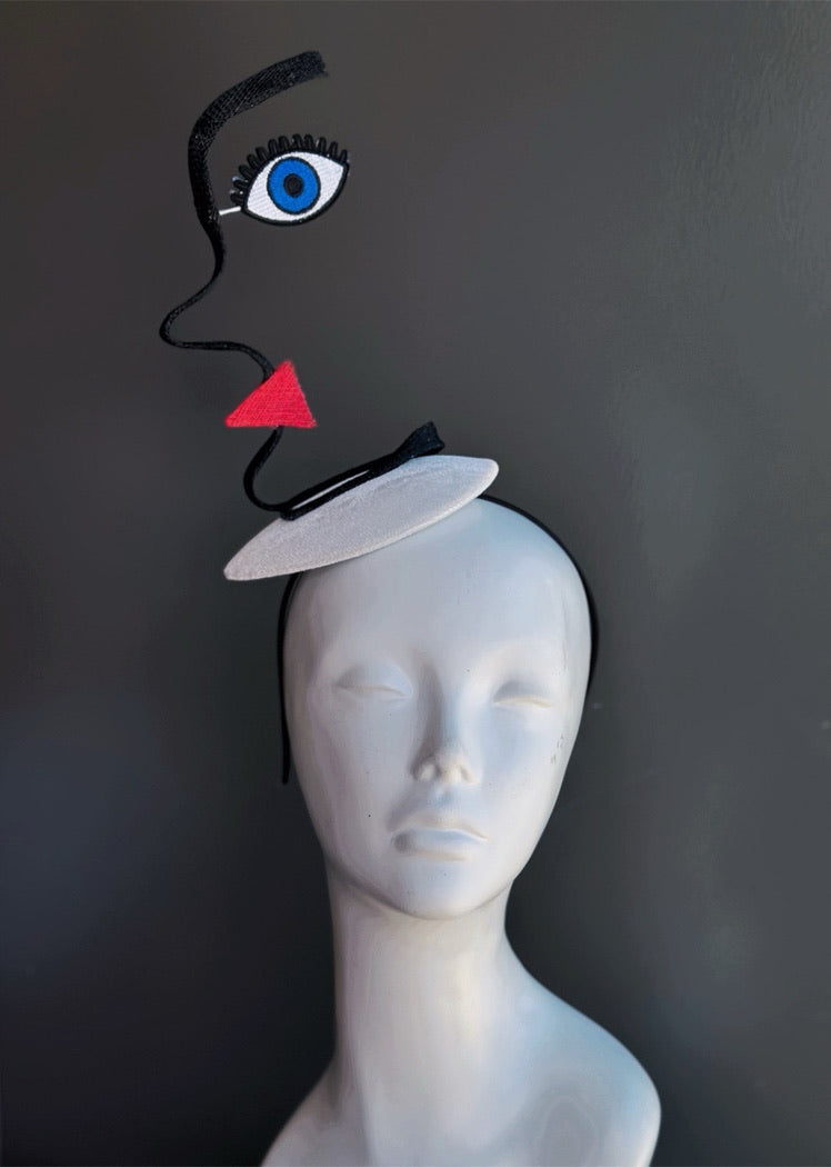 Abstract face fascinator with sculptural black wire silhouette, embroidered blue eye motif, and geometric red lip accent on white fascinator base