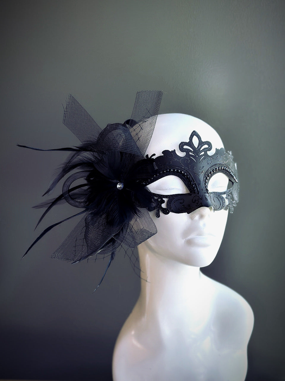 Black feather masquerade mask with a bow accent and intricate filigree displayed on a mannequin head.

