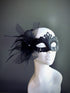Black feather masquerade mask with a bow accent and intricate filigree displayed on a mannequin head.

