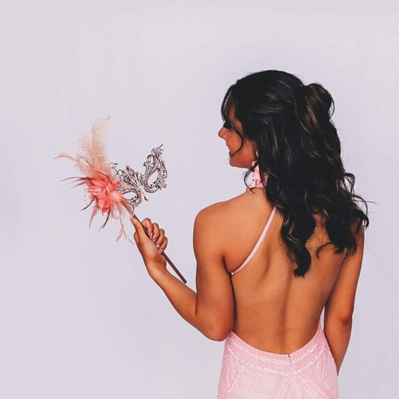 Elegant blush pink masquerade mask with filigree base, floral centerpiece, and soft feathers for prom, Venetian balls, and carnival parties.