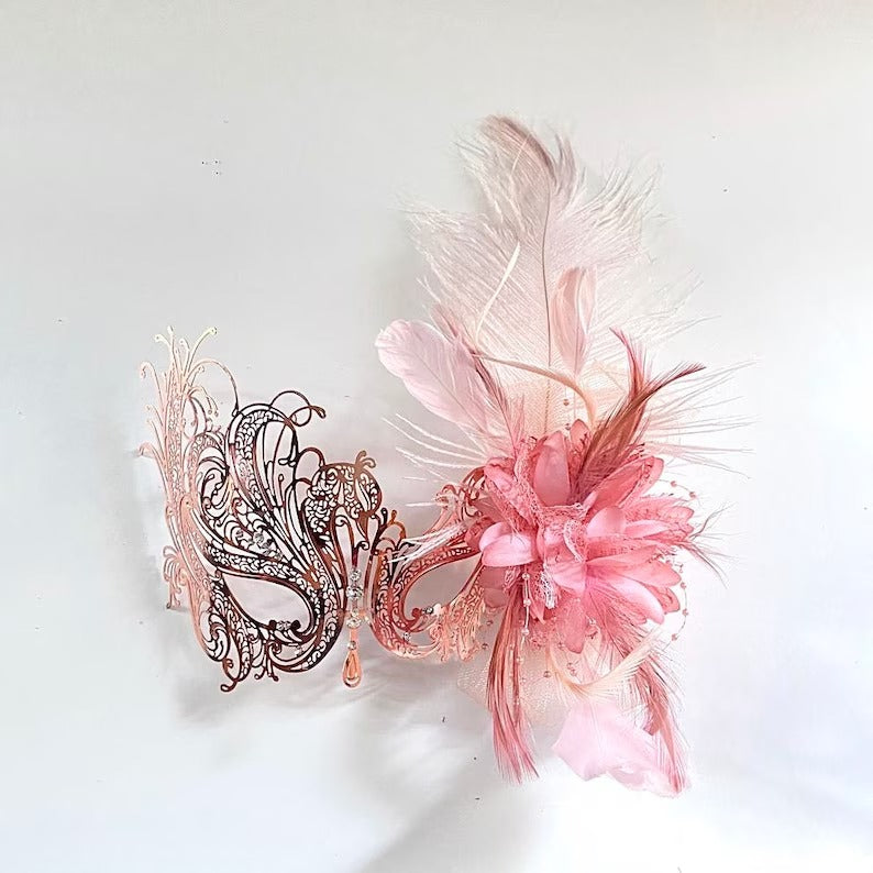 Elegant blush pink masquerade mask with filigree base, floral centerpiece, and soft feathers for prom, Venetian balls, and carnival parties.