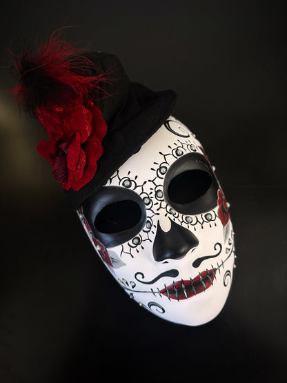 Day of the Dead mask with black and white sugar skull design, burgundy flower, and feather accents, perfect for Día de los Muertos celebrations.
