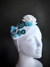 White fascinator hat with a white rose, blue florals, and intricate beaded embellishments.