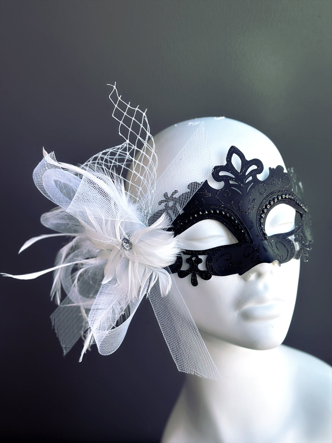 Black metal masquerade mask with white feathers, netting, and floral lace details on mannequin

