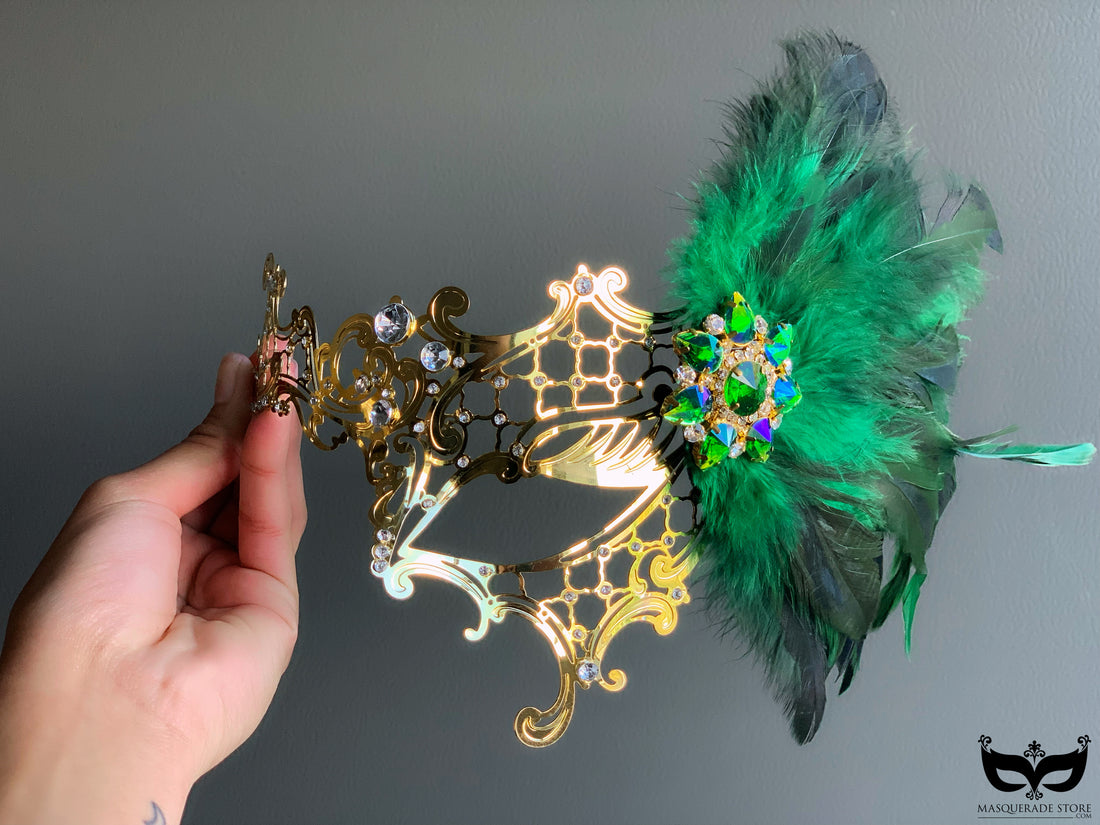 Gold metal filigree mask adorned with green feathers and a multicolored brooch, ideal for luxury events and masquerade balls.