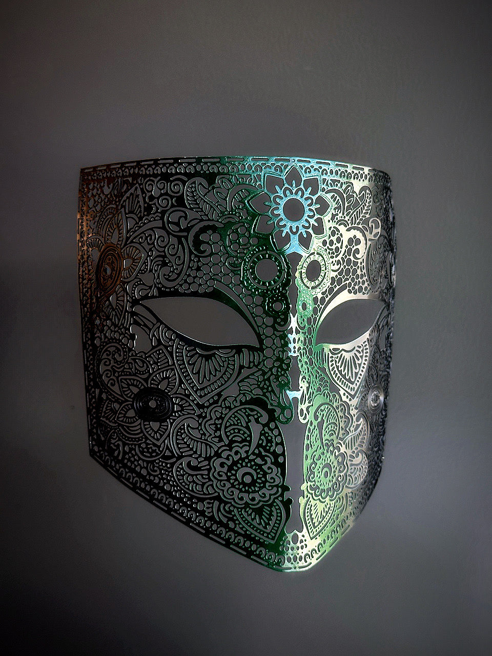 Laser-cut metal mask with an ombre effect, fading from emerald green in the center to black on the sides, featuring intricate filigree detailing.

