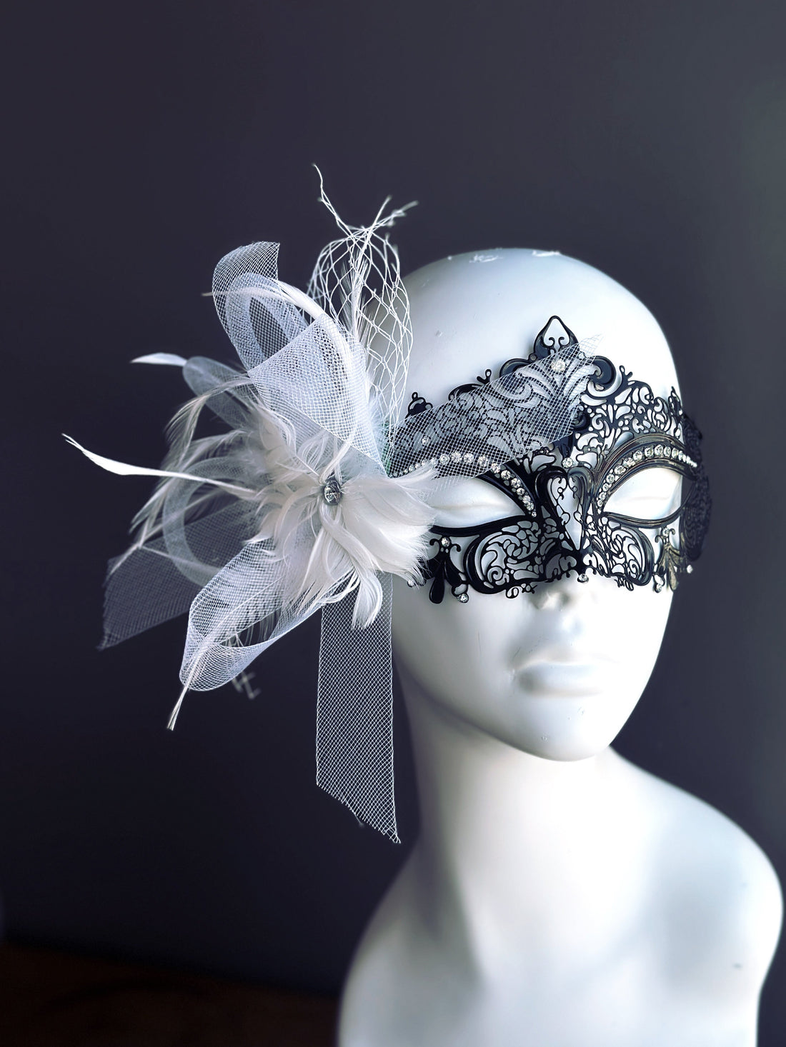 Black metal masquerade mask with rhinestones and white feathers on mannequin
