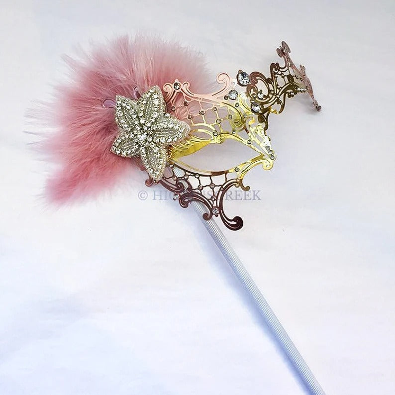 Handheld masquerade mask in pink and gold with rhinestone flower, blush feathers, and elegant stick design for proms, balls, and parties.