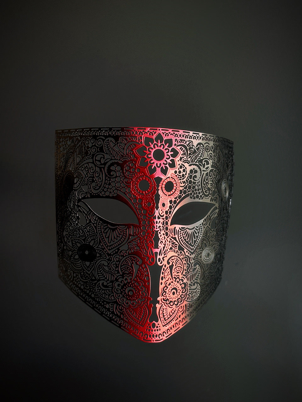Laser-cut metal mask with an ombre gradient from deep crimson red in the center to black edges, featuring intricate filigree detailing.

