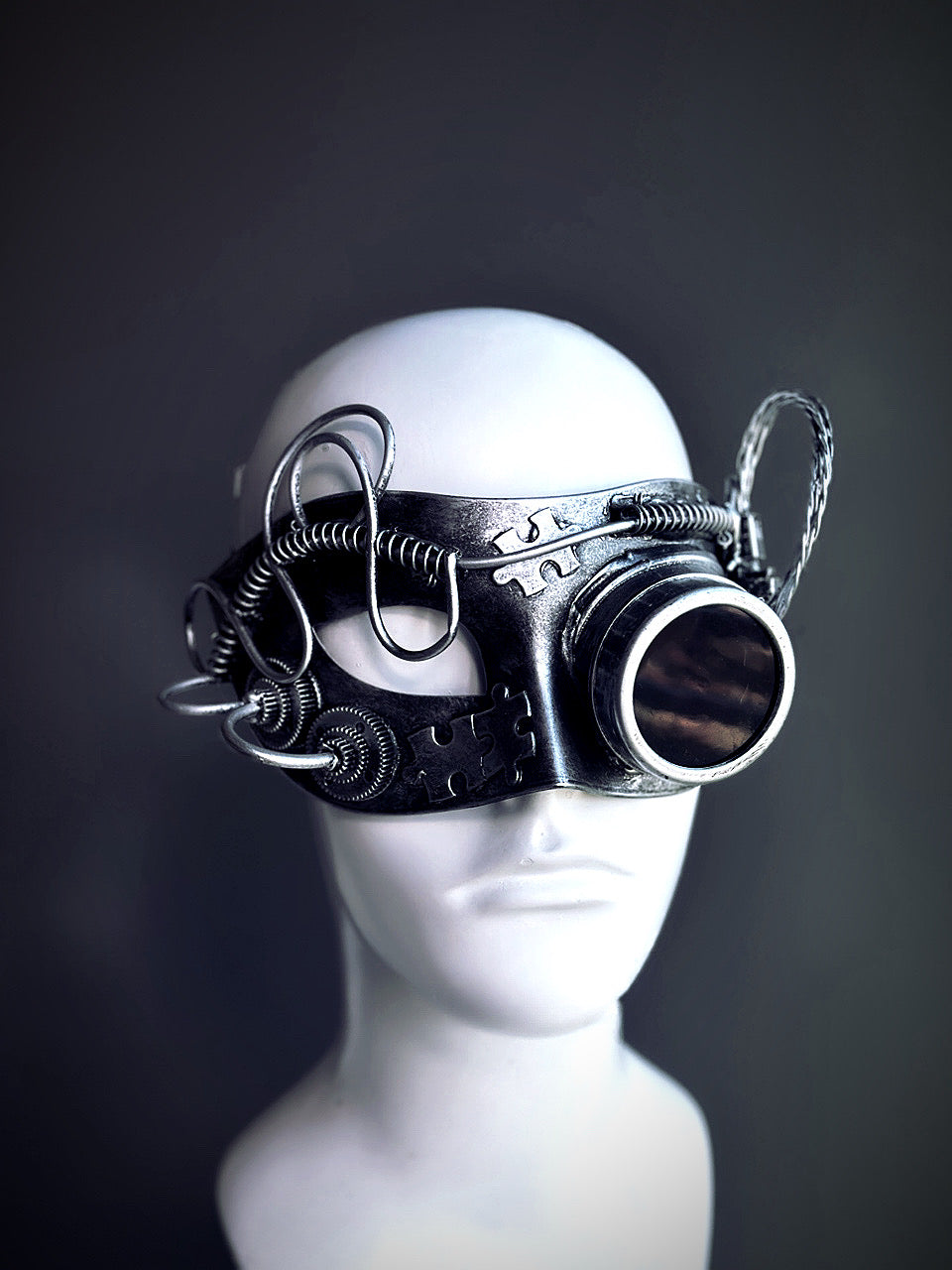 Silver steampunk masquerade mask with a monocular lens, coiled wire, and mechanical details displayed on a mannequin head.